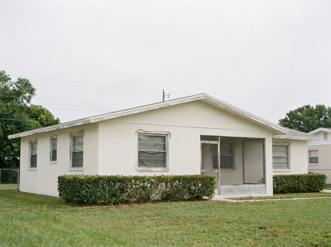 The same illustrated Florida block home under a flat gray sky with old faded aluminum frame windows, dark screens, and stained caulk lines